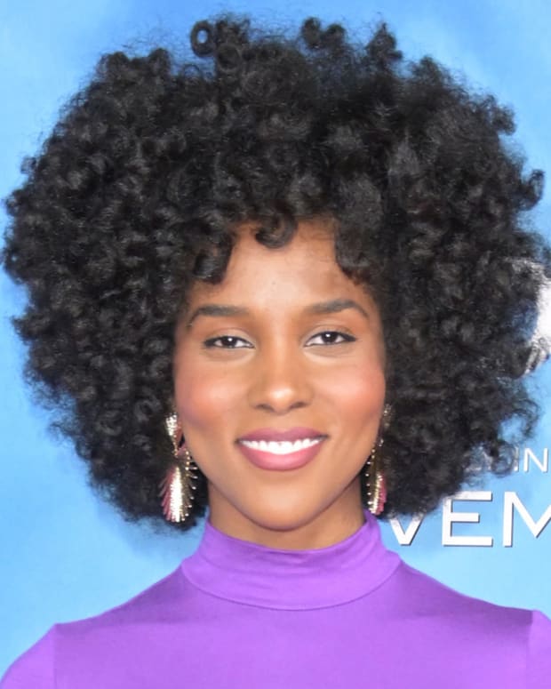Drew Dorsey poses in a purple high-neck silk dress in front of a blue backdrop.
